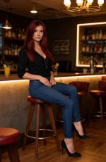 Amber Murphy, the Candy AI generated character, sits confidently on a bar stool in a softly lit lounge, dressed in fitted jeans and heels against a stylish cocktail bar backdrop.